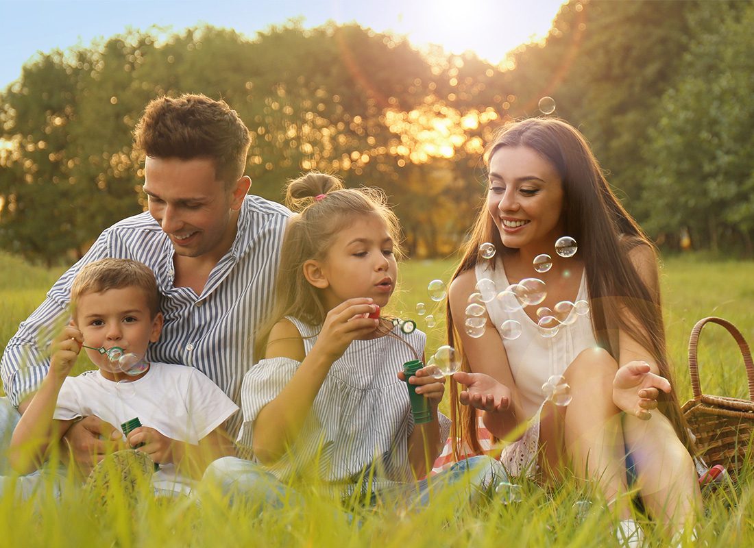 Personal Insurance - Family Sitting in Grass in a Field While Blowing Bubbles and Smiling While the Sunsets Behind the Trees on a Nice Day